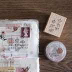 Packaging with floral design, stamp, wooden stamp block, and 'paper cranes' container on a wooden surface.