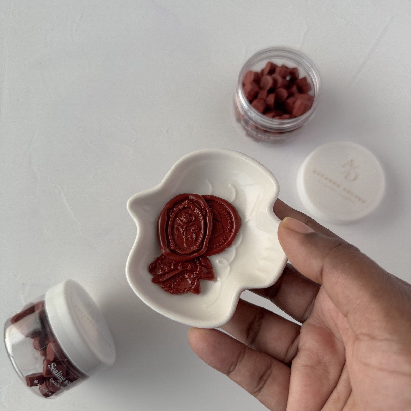 Hand holding a white container with red objects, surrounded by additional containers on a light background