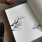 Open notebook with black ink tree drawings on a wooden surface with a pen.