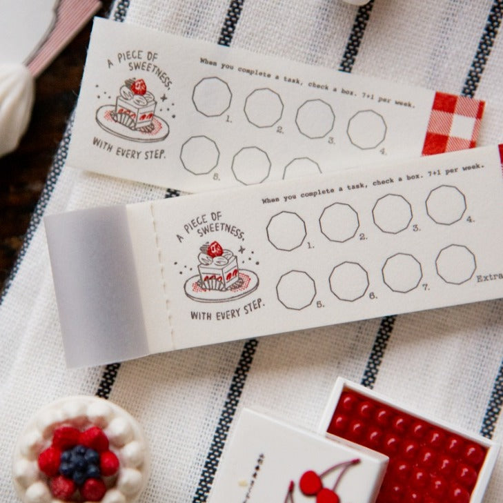 Two reward charts on a checkered tablecloth with a small cake and red building block in the foreground.