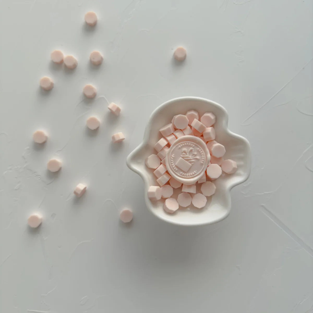 Minimal flat lay of soft pink wax seal beads scattered around a small ceramic dish holding a decorative wax seal stamp, on a clean neutral background.