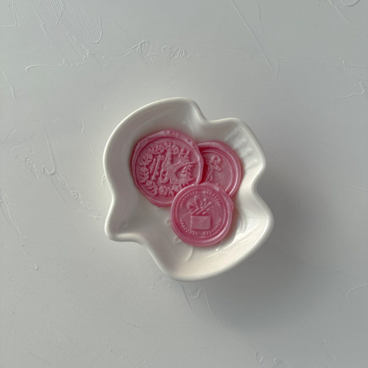 Pearly pink Pink Pearl sealing wax beads and a finished seal gathered in a white bowl, flat lay on soft grey
