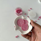 Pink wax seals in a white dish held by a hand with a container of additional seals in the background.