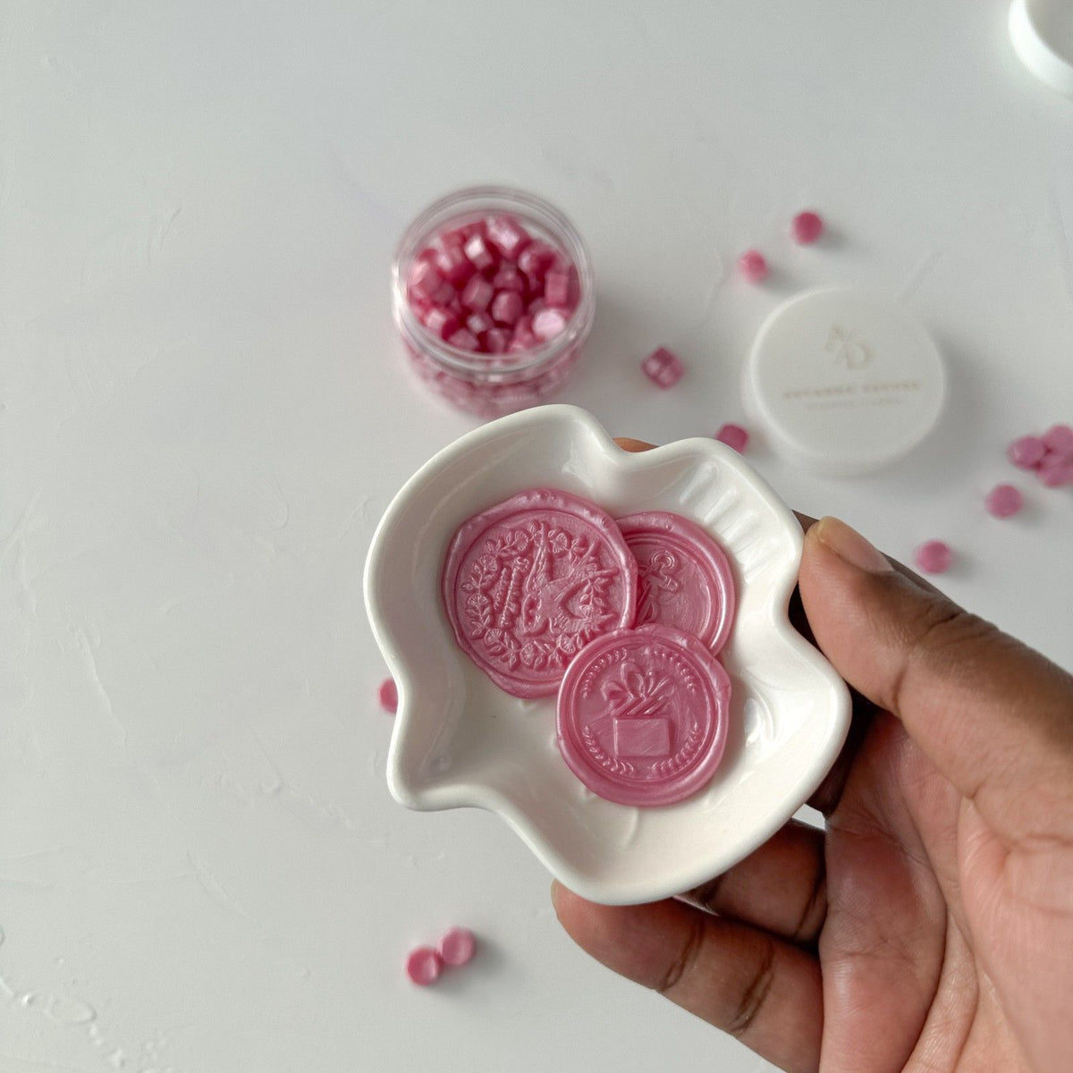 Hand holding a white dish of Pink Pearl Wax Beads seals with the container of loose beads in the background