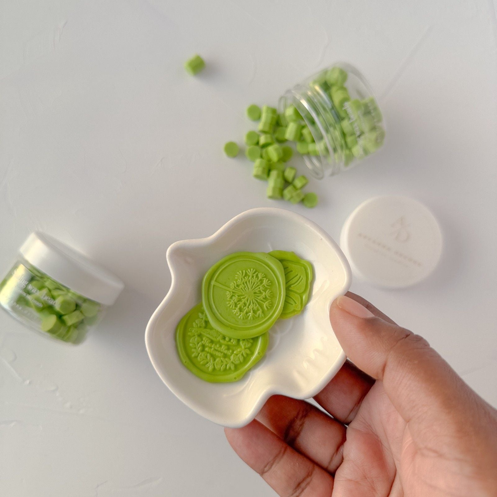 Hand holding a white dish with green cookie-like shapes, surrounded by more green shapes on a white background.