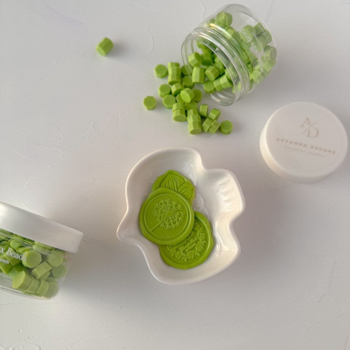 Pistachio Wax Beads in a jar beside a dish of round melted seals on a light grey backdrop