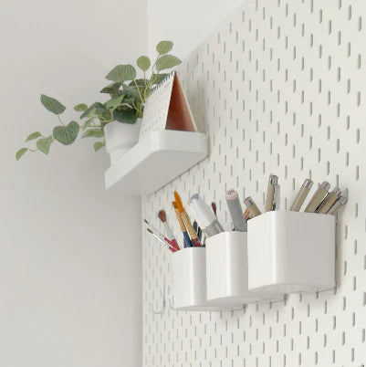 Minimal white pegboard wall organizer with mounted shelf, plant decor, and pen holders storing stationery items