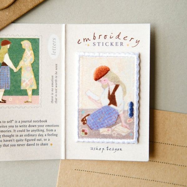 An open notebook or journal page featuring a stitched embroidery sticker with a woman in a red beret and blue skirt, alongside printed text and another small embroidered illustration. The sticker is placed on a beige surface with a kraft envelope corner visible.
