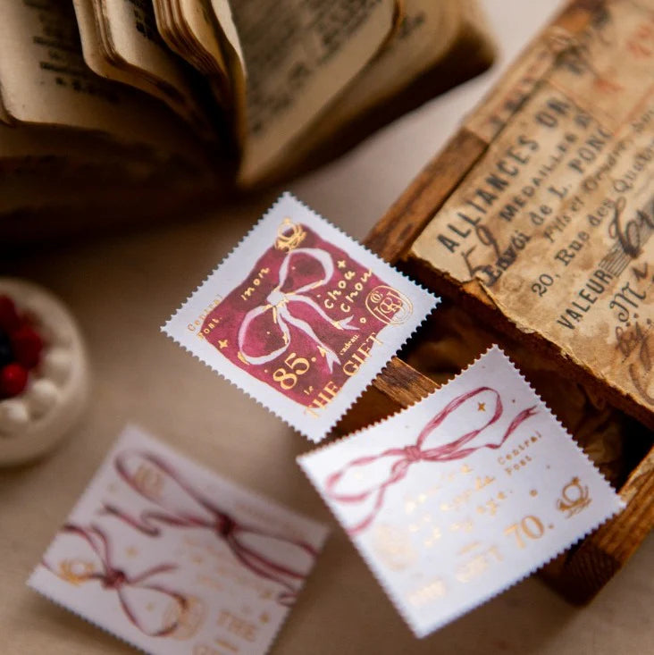 Close-up of vintage-style bow stamp stickers with gold foil details placed beside an old book and paper ephemera on a warm-toned desk.