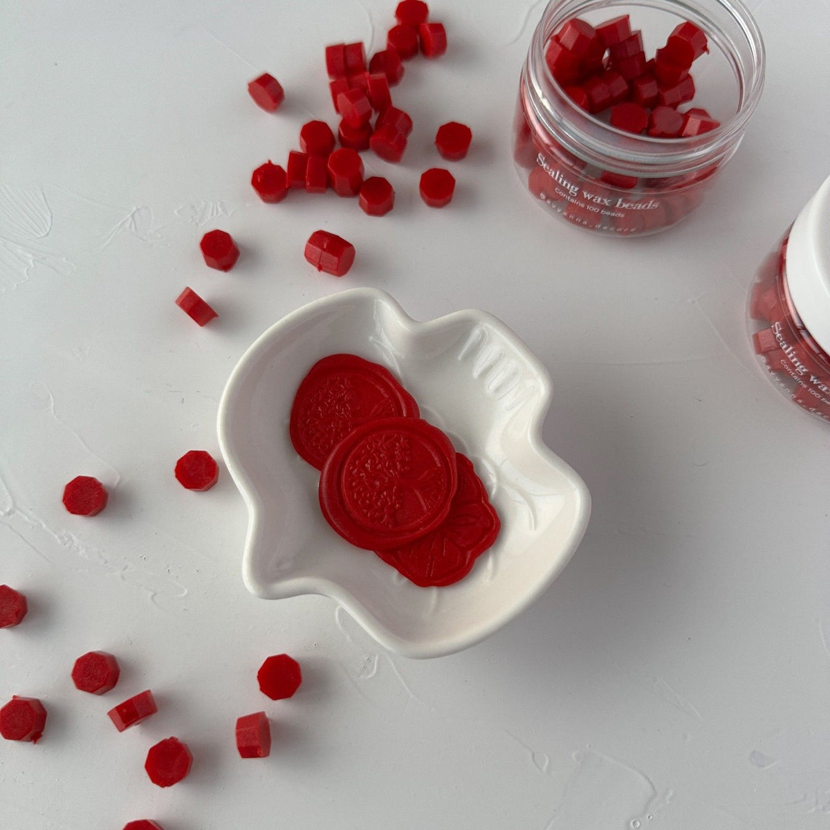Vibrant red Scarlet Wax Beads with melted wax seals in a white dish and jar on a light grey background