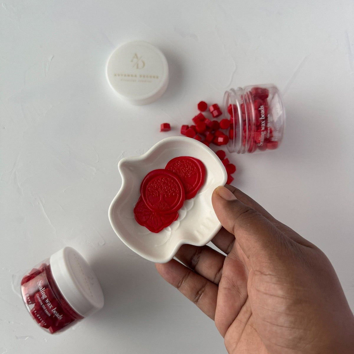 White dish of red Scarlet Wax Beads surrounded by small containers on a light grey backdrop, top view