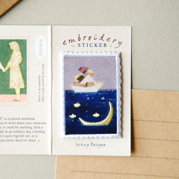 An embroidery sticker displayed on a cream card, with part of another sticker visible to the left. The main sticker shows a stitched scene of a small boat with a figure and books on the water under a pale sky, with a yellow crescent moon and stars below.