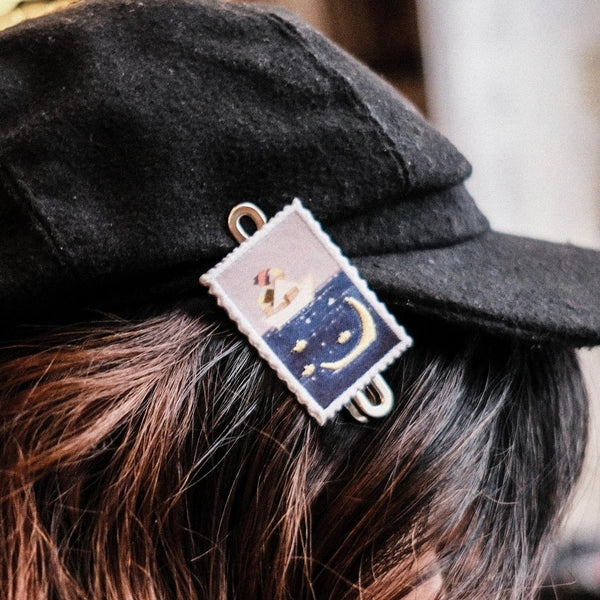 A close-up of a person wearing a black felt cap, with a small rectangular embroidered hair clip holding a strand of brown hair. The clip features a night-sky design with a crescent moon and stars in navy blue and gold, bordered in white.