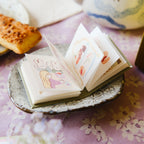 Small open book with cartoon illustrations on a decorative plate with a floral tablecloth.