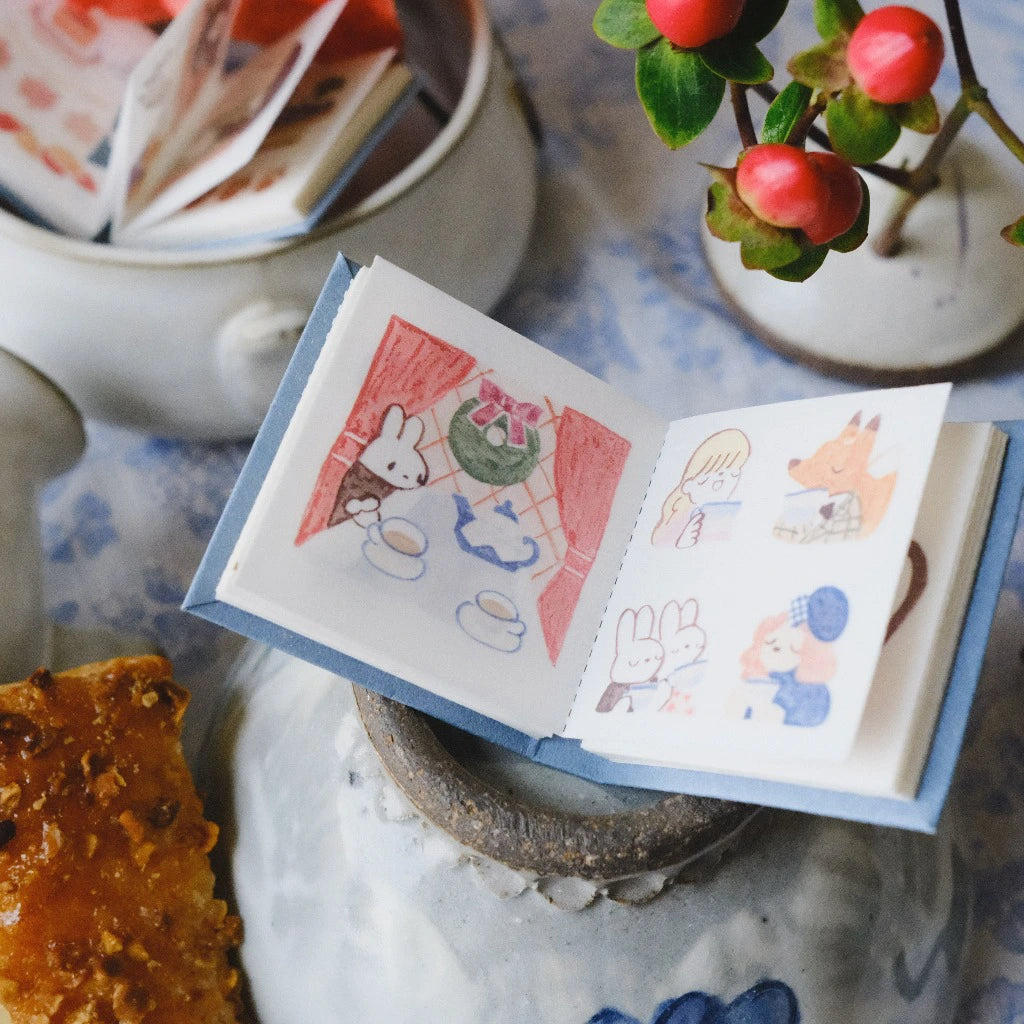 Small open book with illustrations of animals on a decorative surface with a plant and cookies.