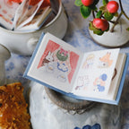 Small open book with illustrations of animals on a decorative surface with a plant and cookies.