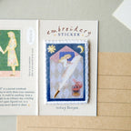 An embroidered patch sticker displayed on a card labeled 'embroidery STICKER'. The patch depicts a stylized needlewoman with a large needle, surrounded by a blue border, with a crescent moon and sun.