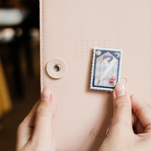 A close-up of a person's hands holding a small rectangular vintage-style illustration card against a pale pink leather journal cover with a snap button.