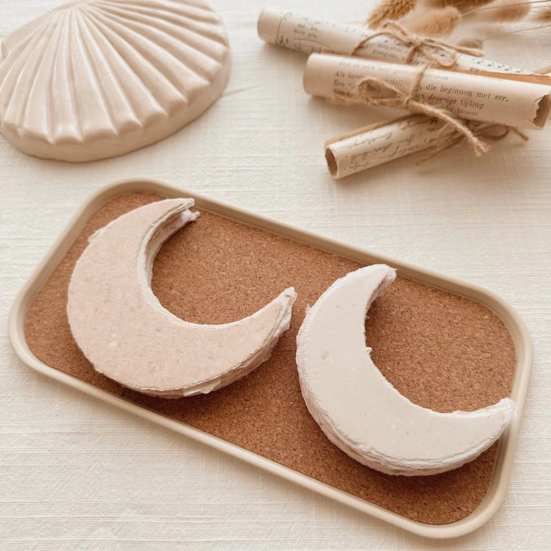 Two Small Moon Handmade Paper crescents on a cork tray with a shell visible behind