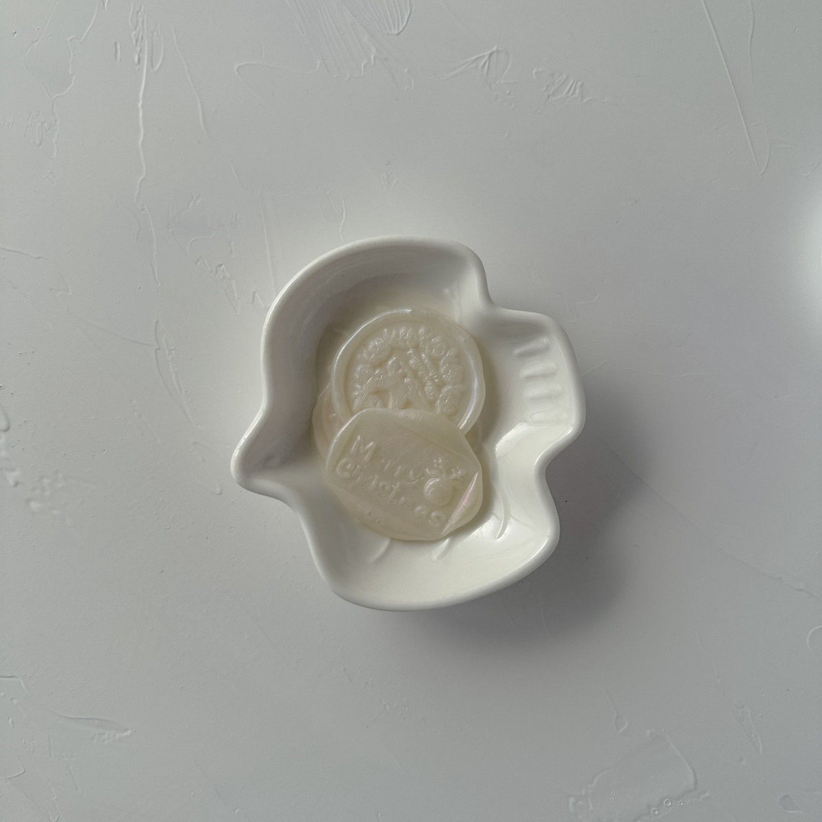 White ceramic dish holding a stamped Soft Glow wax seal emblem, close-up detail on a grey background