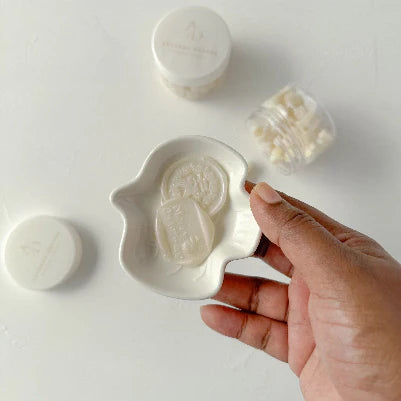 Hand holding a white soap bar with a design, surrounded by other soap products on a light background