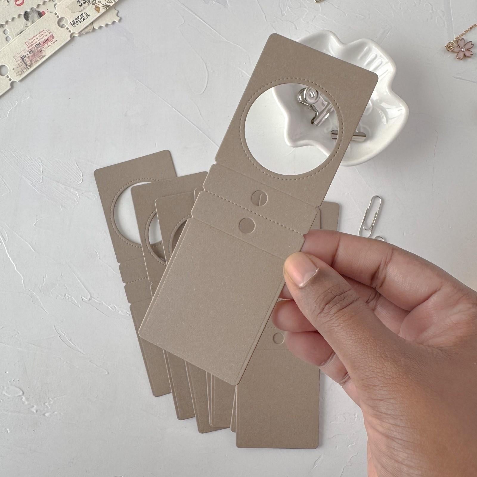 Hand holding a set of beige cardboard earring display cards on a light gray surface.