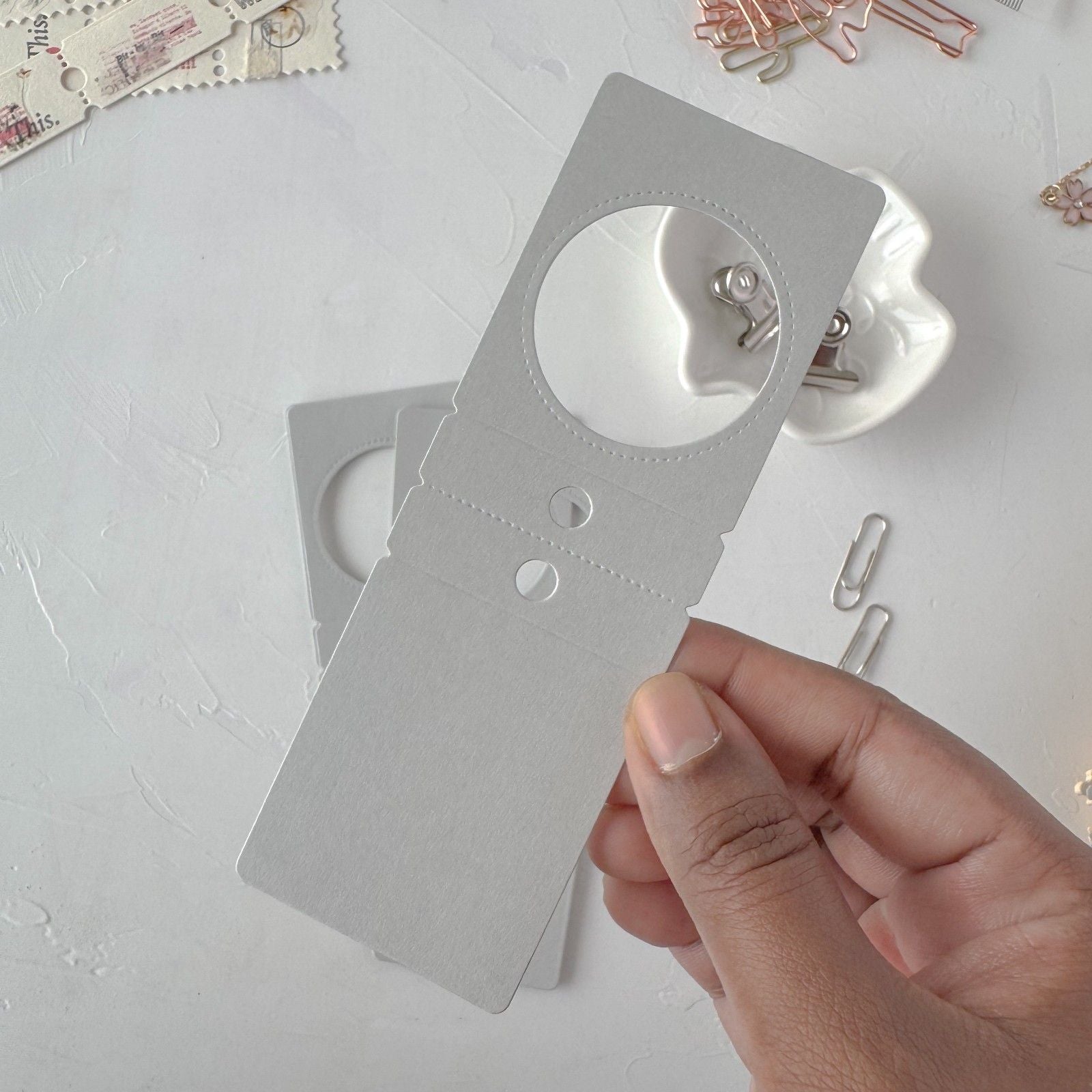 Hand holding a small metal tool on a light gray surface with paper clips.