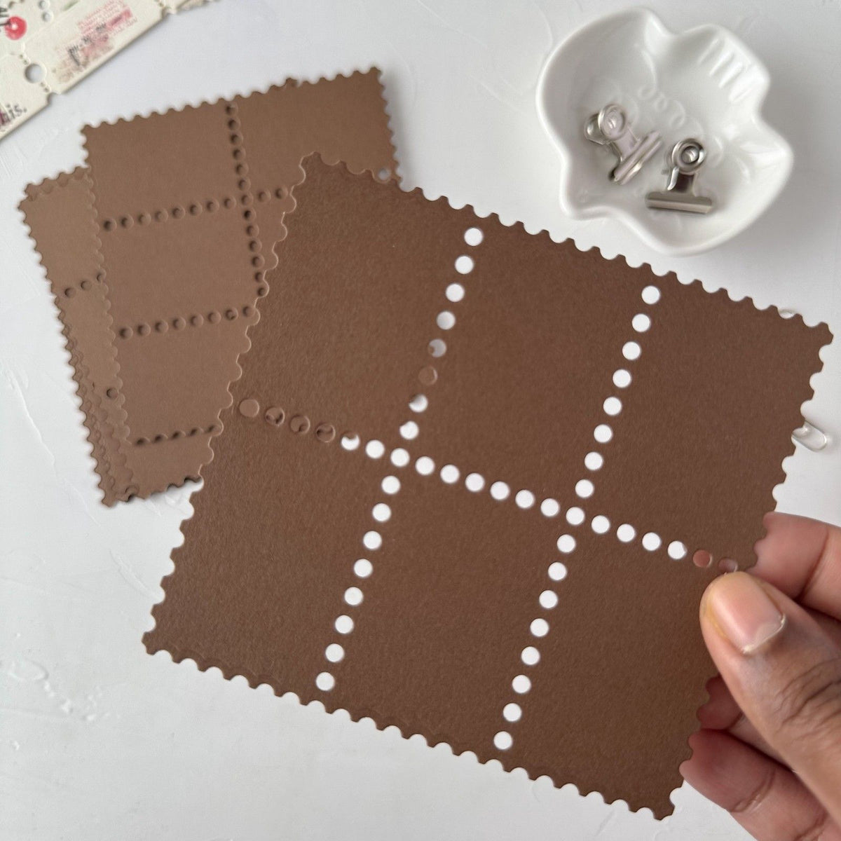 Hand holding a textured brown Stamp Card Vol. 3 square card on a light studio surface