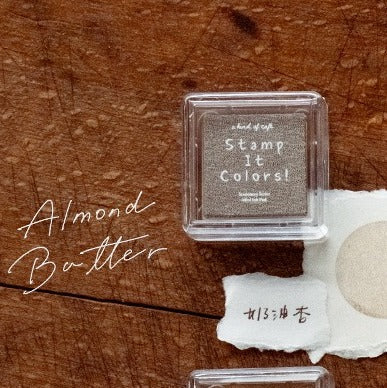 Small container labeled 'Stamp It Colors' on a wooden surface with 'Almond Butter' text.