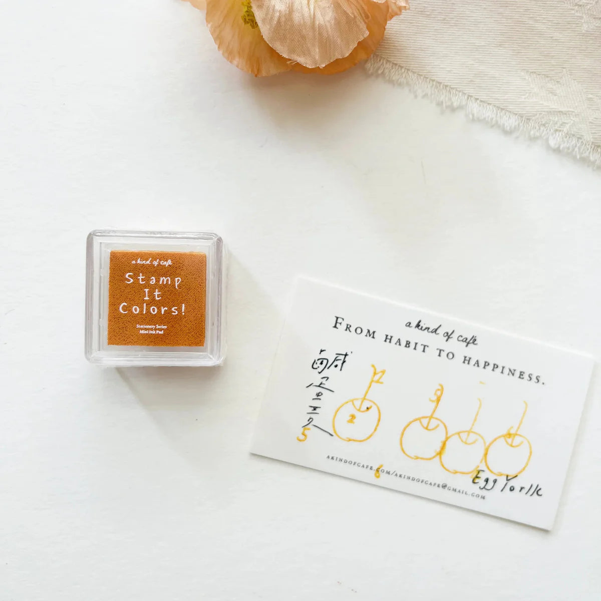 Minimal flat lay of an orange ink pad labeled “Stamp It Colors!” beside a card stamped with simple yellow apple illustrations, on a clean white surface with a soft fabric and flower in the background.