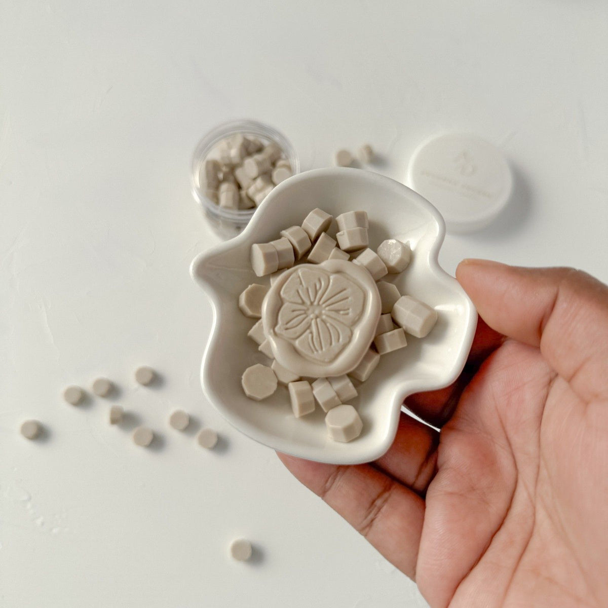 Hand holding a ceramic dish of off-white Steamed Milk Wax Beads showing their small geometric bead shape