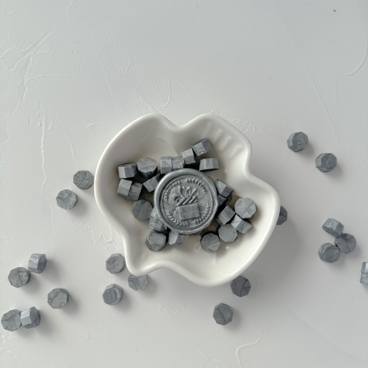 White heart-shaped dish of hexagonal Steel Grey Wax Beads on a light grey studio background, top view