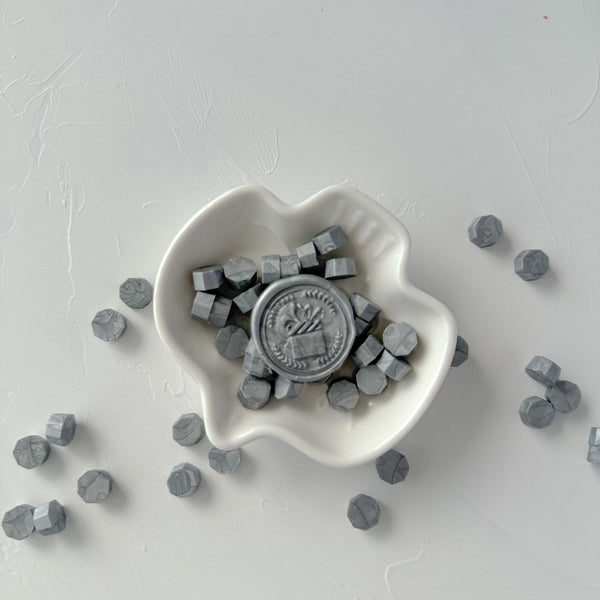 White heart-shaped dish of hexagonal Steel Grey Wax Beads on a light grey studio background, top view