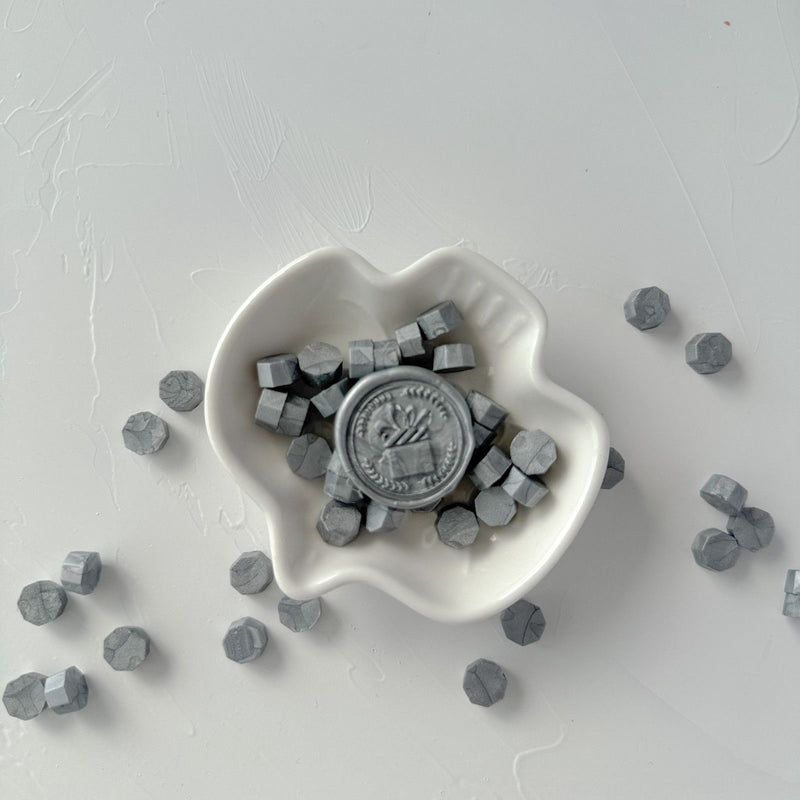 White heart-shaped dish of hexagonal Steel Grey Wax Beads on a light grey studio background, top view