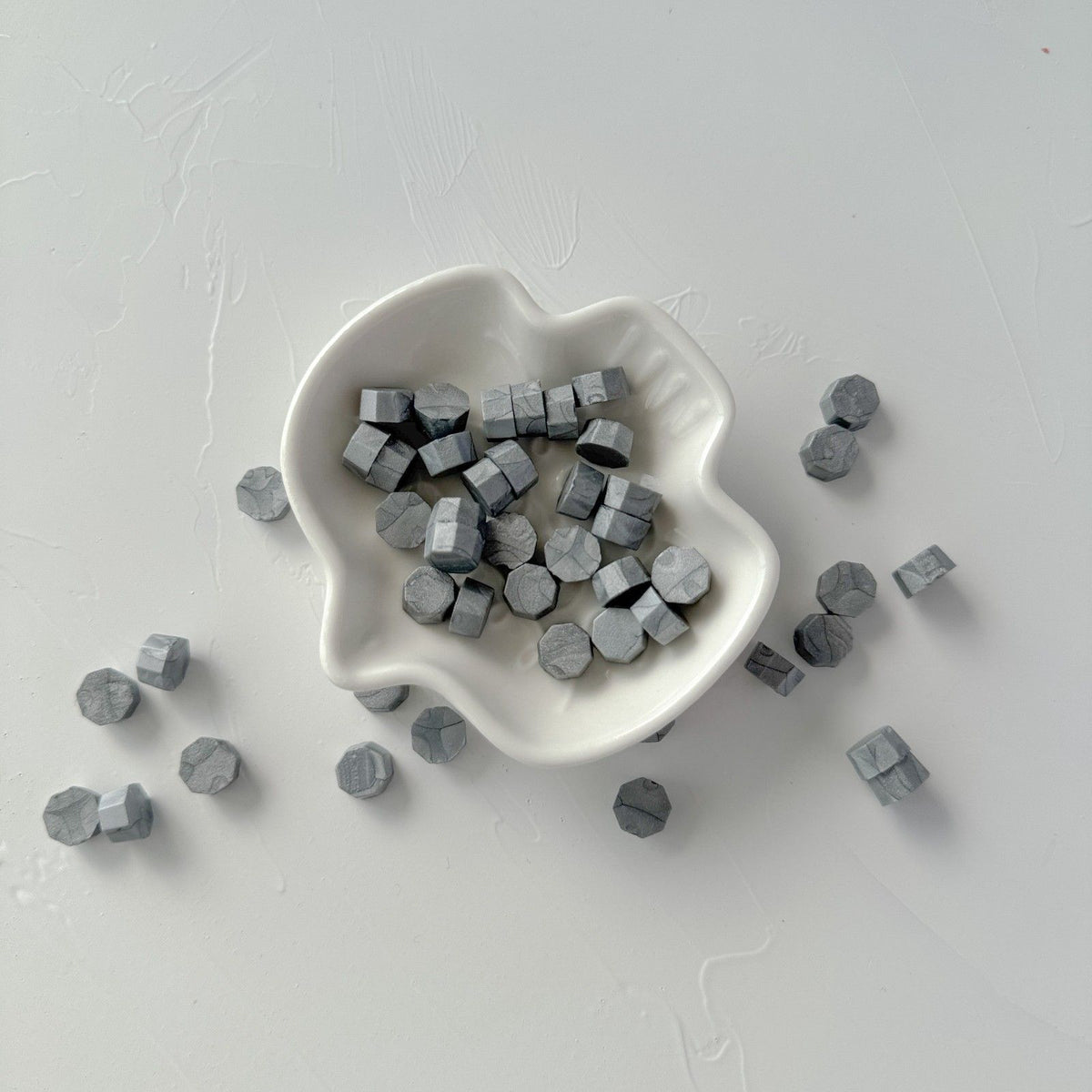 Small white dish filled with metallic Steel Grey sealing wax beads, overhead view on a grey backdrop