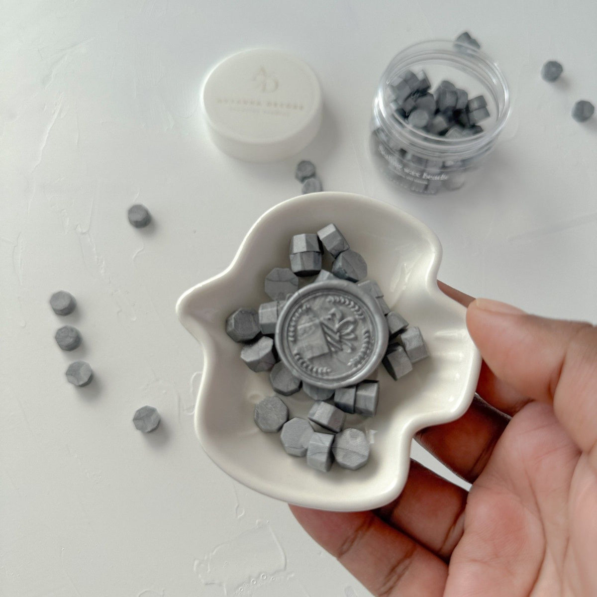 Hand holding a white dish of hexagonal Steel Grey sealing wax beads against a light grey backdrop