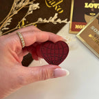 Hand holding a red heart-shaped object with a blurred background featuring chocolate bars.