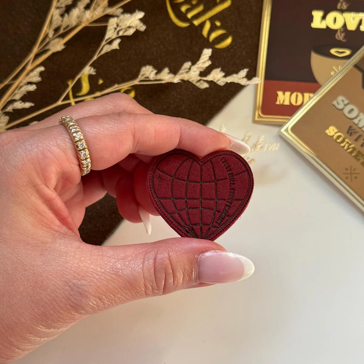 Hand holding a red heart-shaped object with a blurred background featuring chocolate bars.
