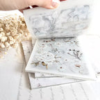 Stack of floral-patterned paper sheets with a hand holding one sheet above them on a light background.