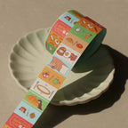 Unrolled strip of breakfast-themed washi tape draped over a plate, showing playful food icons such as pancakes, eggs, bread, and tea.
