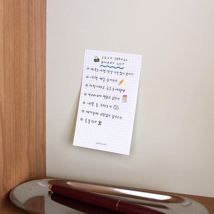 White notepad with handwritten notes attached to a light-colored wall next to a wooden surface.