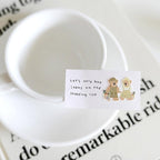 White mug with a small card and two teddy bear illustrations on a newspaper background