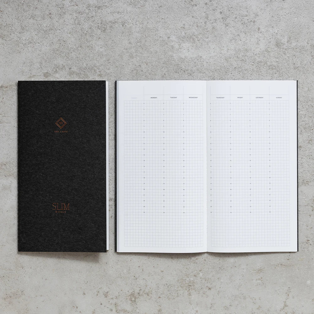 Take A Note Slim Weekly Planner closed in black beside an open grid spread on concrete