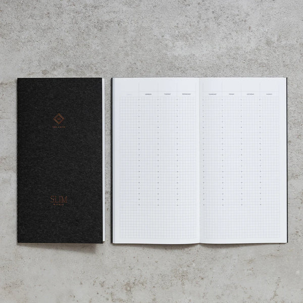 Take A Note Slim Weekly Planner closed in black beside an open grid spread on concrete