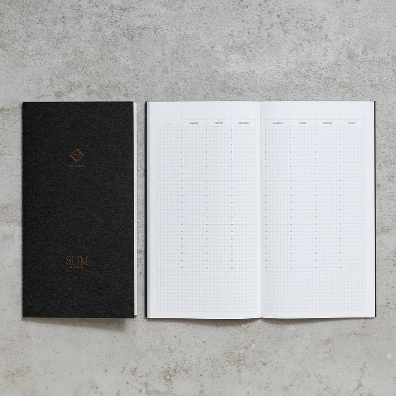 Take A Note Slim Weekly Planner closed in black beside an open grid spread on concrete