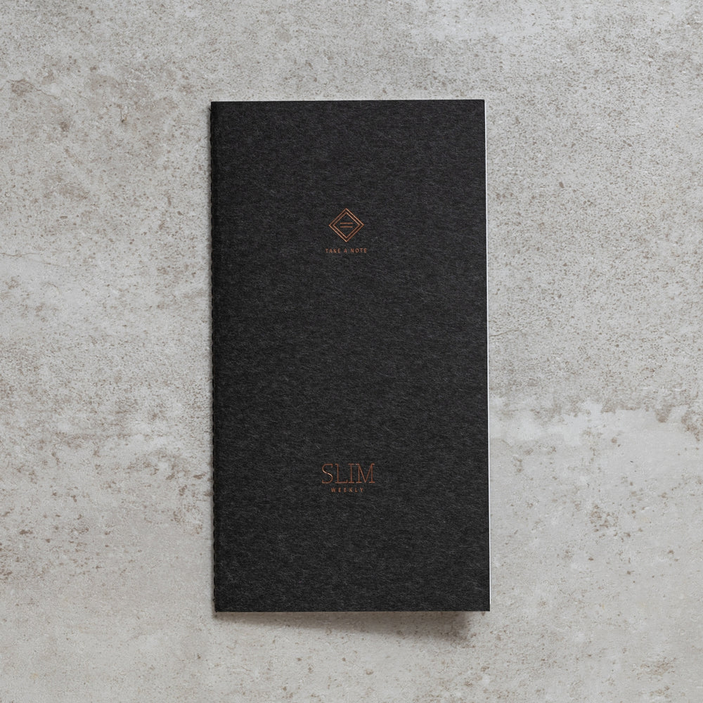 Black Take A Note Slim Weekly Planner with SLIM branding on a textured grey surface