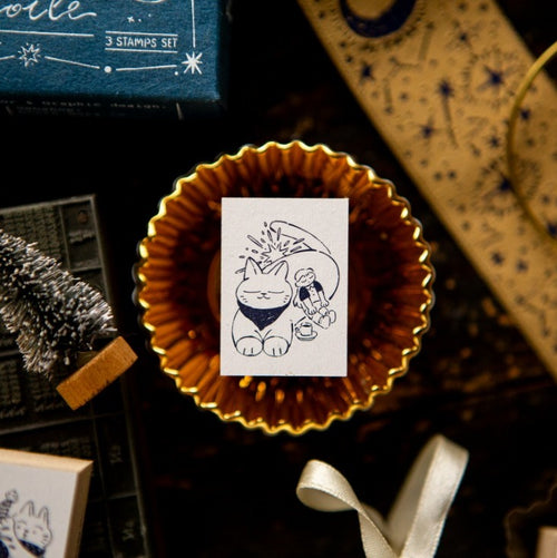 Small white card with a black and white illustration of a cat and a person in a blue dress, placed on a gold circular object.