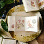 Small open book with floral and animal illustrations on a rustic surface.