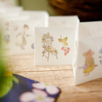 Small square card with illustrations of a chair, butterfly, and flowers on a wooden surface.