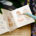 Illustrated book with a girl and butterfly design, held open by a pen.